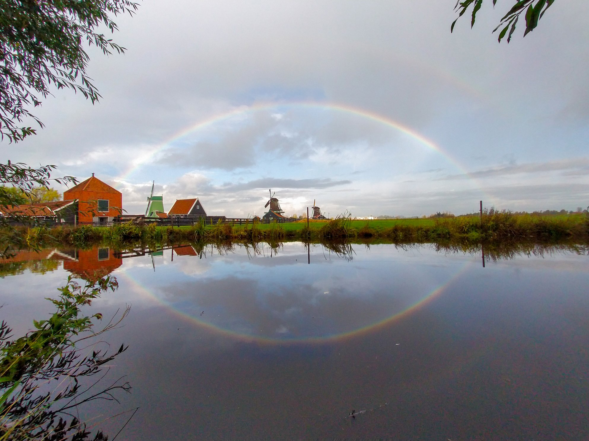 Circular rainbow over a lake with a building, windmill and field in the background